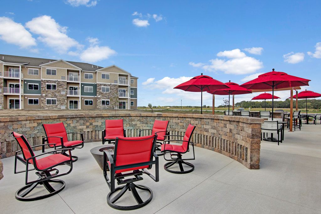 A community patio here at Club at Heritage with cushioned seating and a fire pit area. Red chairs and umbrellas add a vibrant touch, while open views and surrounding buildings create a welcoming outdoor gathering space.